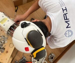 Persona trabajando en la reparación o ensamblaje de maquinaria industrial, usando casco y protección auditiva, con equipo de MRI visible.