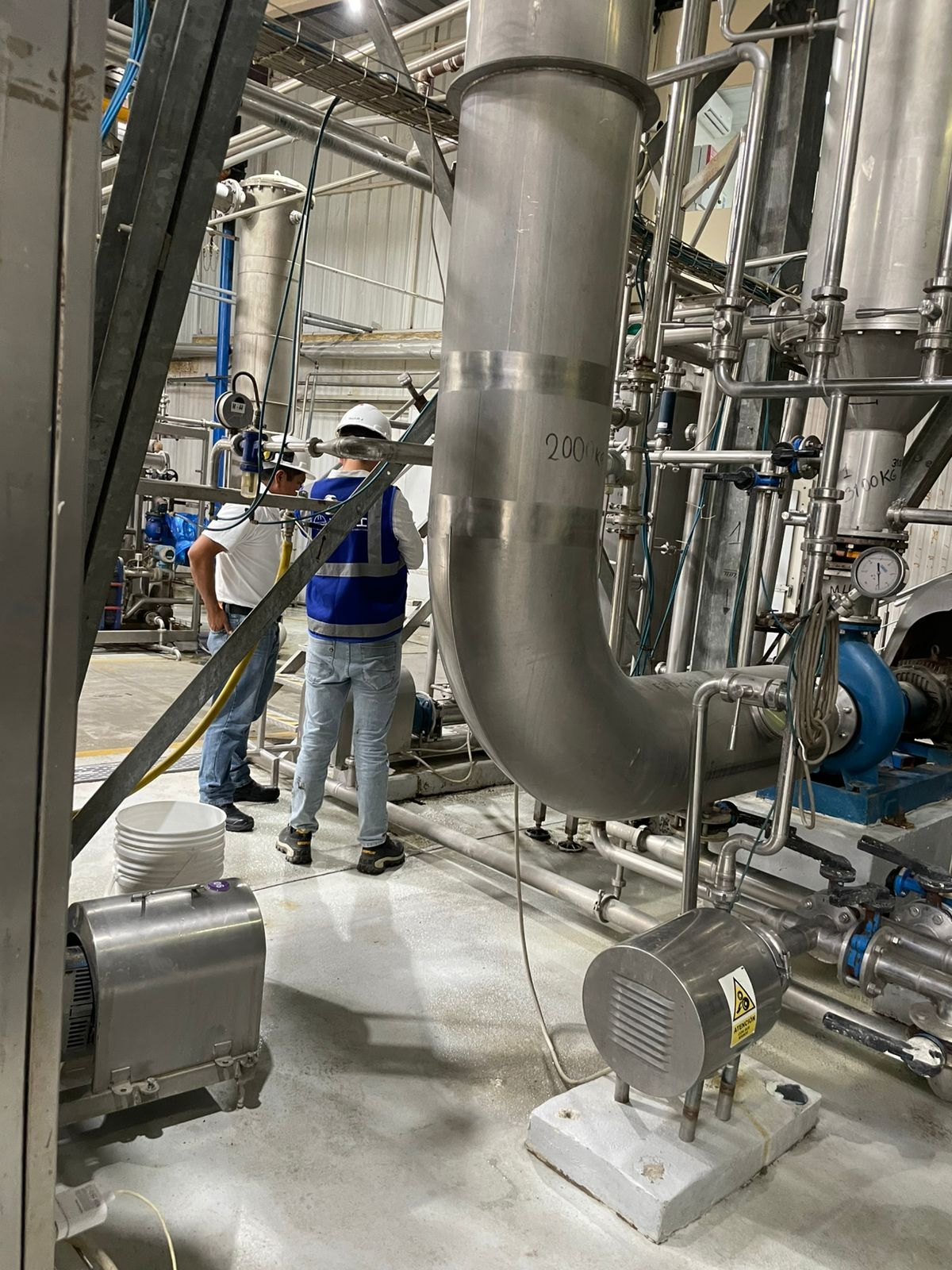 Personal técnico con casco y chaleco realiza inspección en planta industrial, junto a bombas, tuberías de acero inoxidable y equipos de proceso en funcionamiento